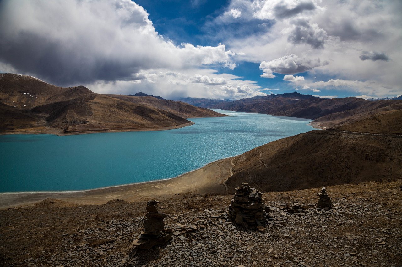 Turquoise Yamdrok Lake under spring clouds in late March, with stone cairns on the pass